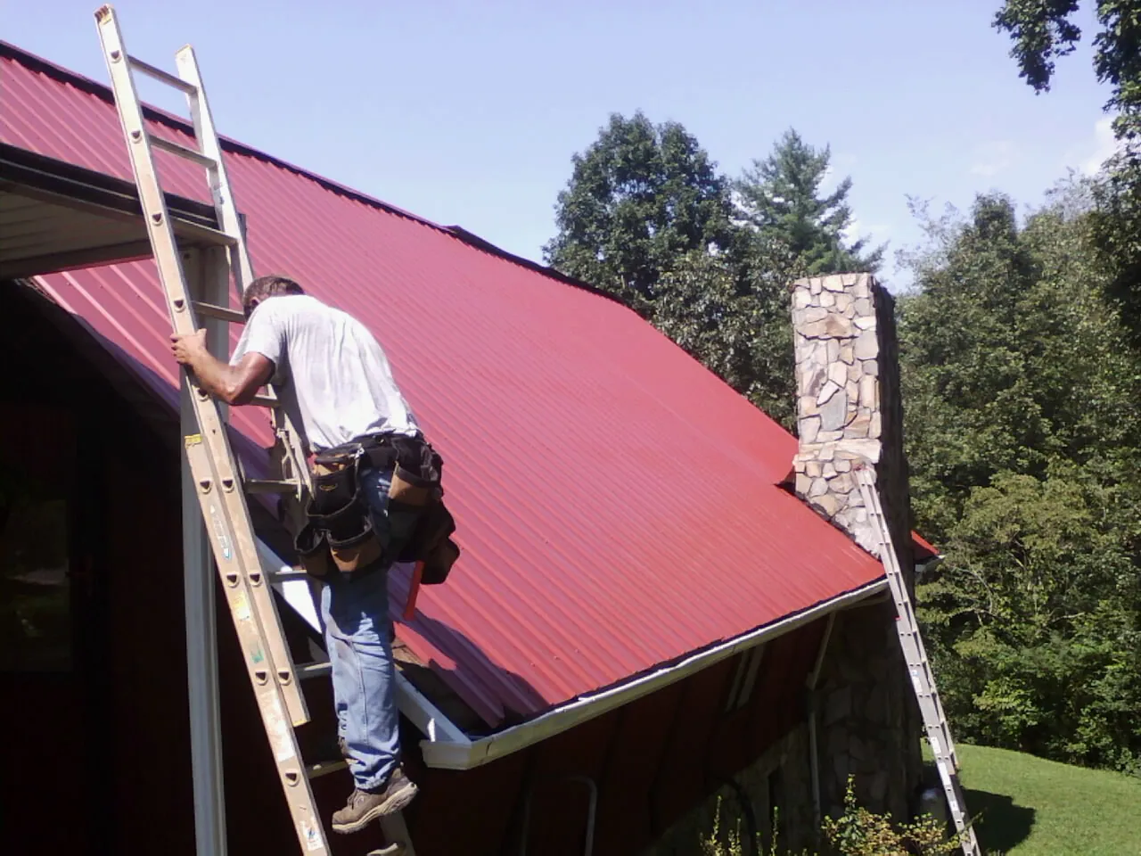 Roofer on a ladder beside a red metal roof and stone chimney — Soffit & Fascia Repair by Roofing Brothers in Jacksontown
